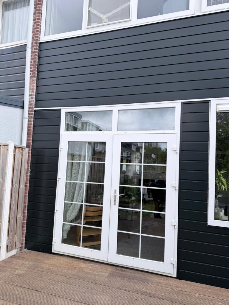 Voor dit project hebben de bewoners gekozen voor Keralit gevelbekleding in een moderne donkere kleurstelling. Keralit is een populaire keuze bij renovaties vanwege de combinatie van duurzaamheid, uitstraling en onderhoudsgemak. In tegenstelling tot houten gevelbekleding hoeft Keralit niet geschilderd te worden. Het materiaal is kleurvast, bestand tegen vocht en goed bestand tegen de Nederlandse weersomstandigheden. Hierdoor blijft de gevel jarenlang strak ogen zonder dat er veel onderhoud nodig is. De gekozen donkere kleur zorgt bovendien voor een moderne uitstraling die mooi contrasteert met de witte kozijnen en deuren aan de achterzijde van de woning.